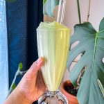 avocado smoothie in a milkshake glass with a glass straw, held in front of some houseplants