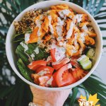 Vegan buffalo chicken in a bowl with quinoa, tomato, cucumber, and romaine lettuce. Palm in the background.