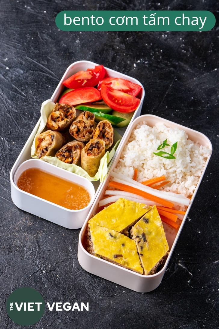Vietnamese bento with two layers, one with rice, egg meatloaf, pickled carrots and daikon, the other with spring rolls and vegetables and sauce