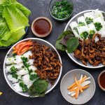 Overhead shot of rice vermicelli noodle bundles on a plate with panfried soy curls, tomato, red mint, with sauce, lettuce, and pickled carrots & daikon