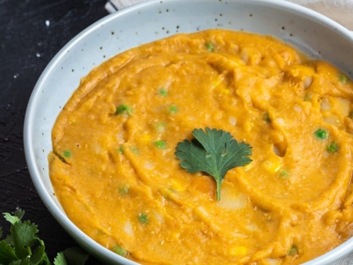 soup in a wide, shallow bowl, garnished with a cilantro leaf on a dark background, surrounded by garlic, a spoon, a tea towel, and cilantro, with "
