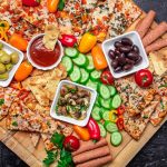 Overhead shot of snack board, three bowls contain black olives, green olives, sauce, and marinated mushrooms, and are surrounded by baked Wholly Veggie Pizza and sliced fresh veggies