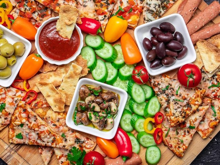 Overhead shot of snack board, three bowls contain black olives, green olives, sauce, and marinated mushrooms, and are surrounded by baked Wholly Veggie Pizza and sliced fresh veggies