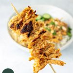 satay on a stick in foreground, with bowl of satay, rice, and green papaya salad in the background