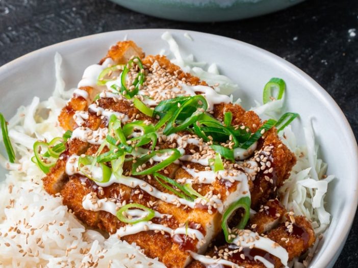 Vegan Tofu Katsu in a bowl drizzled with tonkatsu sauce, vegan mayo, scallions, sesame seeds, on a bed of shredded cabbage and rice