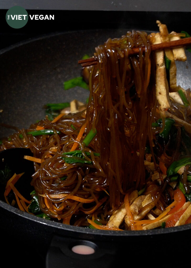 chopsticks pulling up a large amount of sweet potato vermicelli in a wok full of japchae