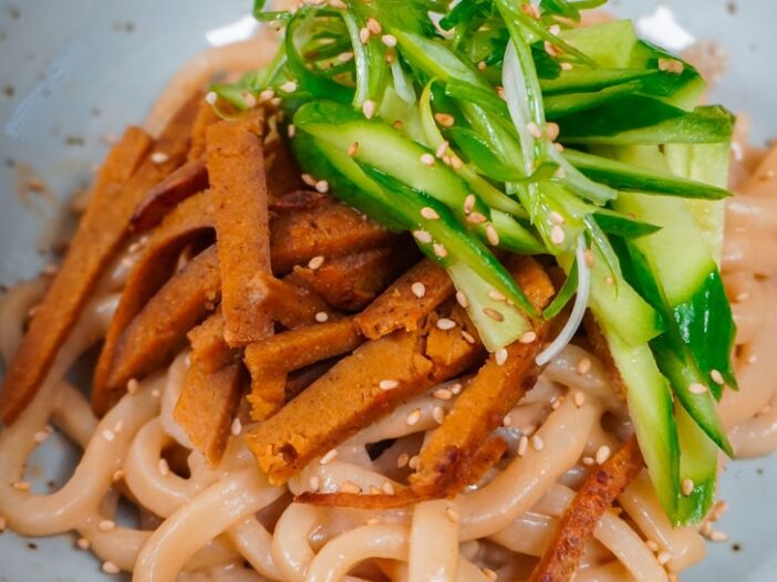 sesame udon topped with sliced seitan, cucumber, and green onion curls